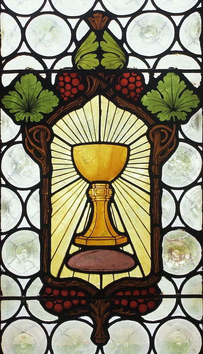 Kirchenfenster in der Sakristei. Zeigt einen goldenen Kelch umrandet von Weinreben und Trauben in der Fensterscheibe